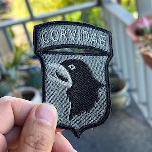 May include: A gray and black embroidered patch with the text "CORVIDAE" and a black bird silhouette.