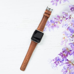 May include: A brown leather watch band with a silver buckle and a black watch face. The watch band is lying on a white wooden surface with lavender flowers in the background.