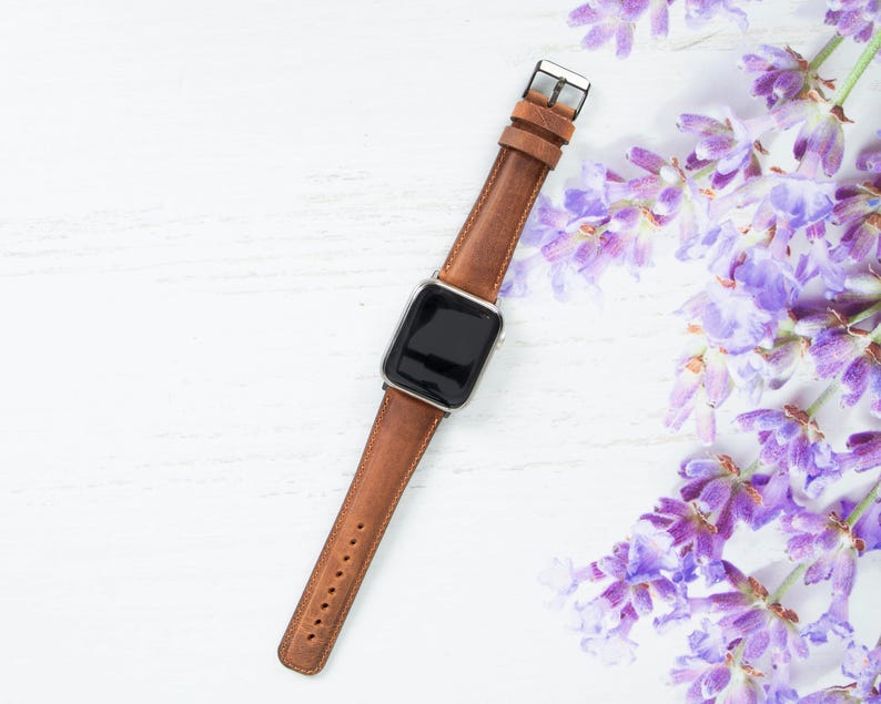 May include: A brown leather watch band with a silver buckle and a black watch face. The watch band is lying on a white wooden surface with lavender flowers in the background.