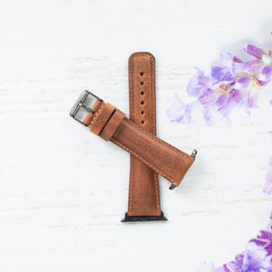 May include: A brown leather watch band with a silver buckle. The band is on a white surface with purple flowers in the background.