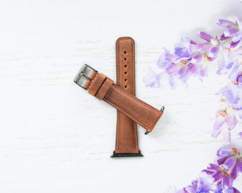 May include: A brown leather watch band with a silver buckle. The band is on a white surface with purple flowers in the background.