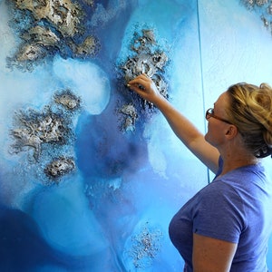May include: A woman is adding finishing touches to a large abstract painting with a textured, blue and silver surface. The painting is on a canvas and appears to be a landscape.