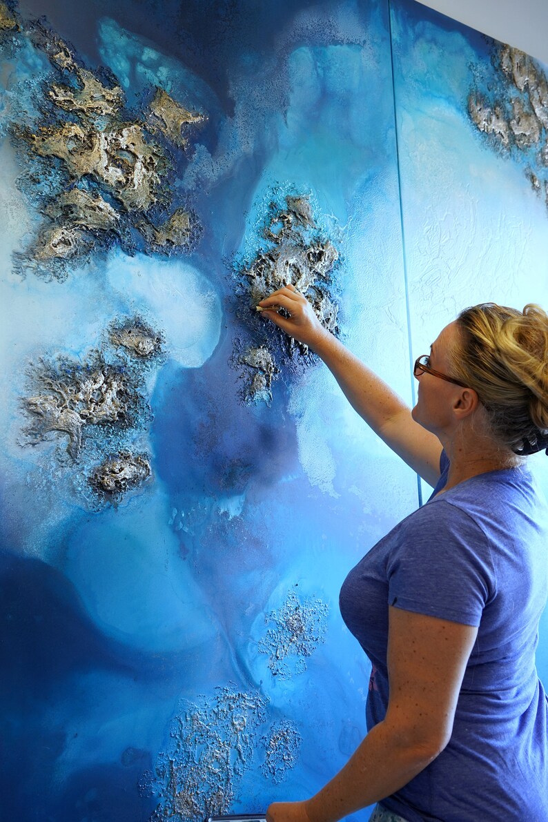 May include: A woman is adding finishing touches to a large abstract painting with a textured, blue and silver surface. The painting is on a canvas and appears to be a landscape.