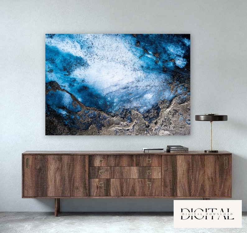 May include: Abstract art print featuring a blue and white watercolor-like design with gold accents. The print is displayed on a wooden dresser with a lamp.