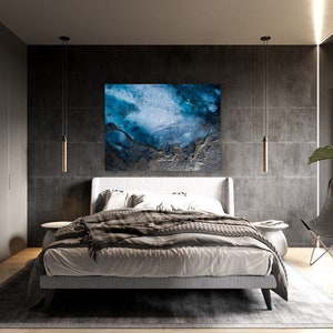 May include: A bedroom with a modern design featuring a gray and white bed with a gray blanket and a blue abstract painting on the wall. There are two pendant lights hanging from the ceiling and a black chair with a plant in a pot next to it.