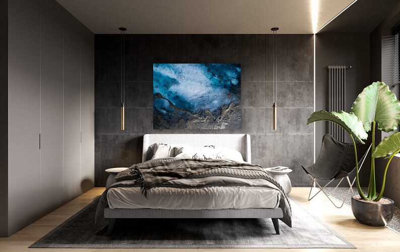 May include: A bedroom with a modern design featuring a gray and white bed with a gray blanket and a blue abstract painting on the wall. There are two pendant lights hanging from the ceiling and a black chair with a plant in a pot next to it.
