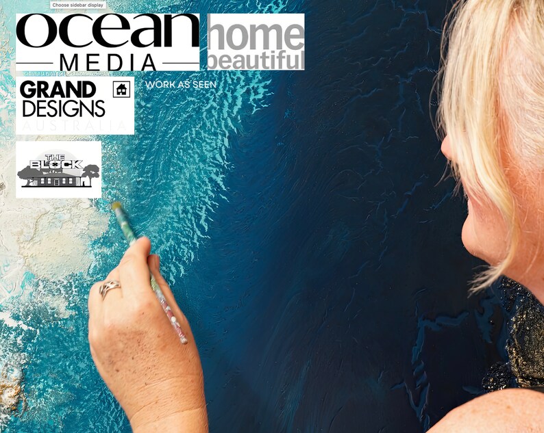 May include: A woman paints a blue and white abstract painting with a brush. The painting has a textured surface and resembles water. The text "ocean home beautiful" and "grand designs" is visible in the upper left corner of the image.