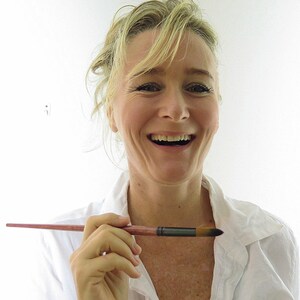 May include: A woman with blonde hair is smiling and holding a paintbrush. She is wearing a white shirt.
