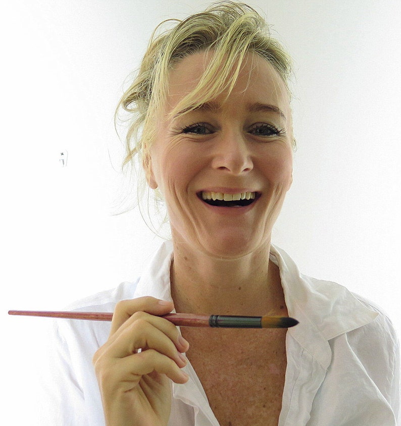 May include: A woman with blonde hair is smiling and holding a paintbrush. She is wearing a white shirt.
