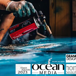 May include: A person using a red and black butane torch to heat a blue surface. The torch is labeled "Trade name" and "Butane Power Gas 2200". The surface is a large piece of art with a blue and white abstract design. The text "GRAND DESIGNS" is visible in the lower right corner of the image. The text "WORK AS SEEN" is visible below the text "GRAND DESIGNS". The text "ocean MEDIA" is visible in the lower center of the image. The text "TIMES SQUARE 2023" is visible in the lower left corner of the image. The text "home beautiful" is visible in the lower right corner of the image.