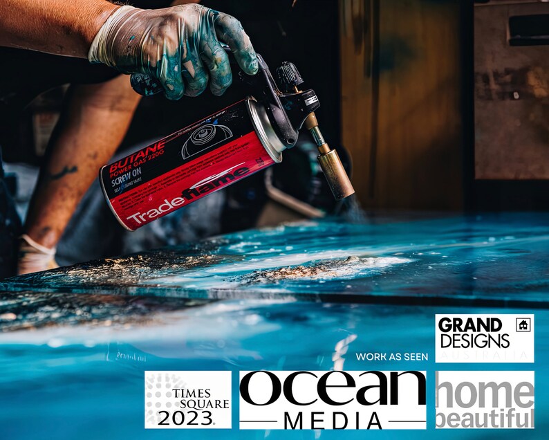 May include: A person using a red and black butane torch to heat a blue surface. The torch is labeled "Trade name" and "Butane Power Gas 2200". The surface is a large piece of art with a blue and white abstract design. The text "GRAND DESIGNS" is visible in the lower right corner of the image. The text "WORK AS SEEN" is visible below the text "GRAND DESIGNS". The text "ocean MEDIA" is visible in the lower center of the image. The text "TIMES SQUARE 2023" is visible in the lower left corner of the image. The text "home beautiful" is visible in the lower right corner of the image.