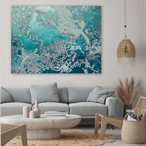 Op de afbeelding: Abstract schilderij in teal en witte kleuren. Het schilderij heeft een textuur, marmerlook en hangt aan een witte muur boven een grijze bank.