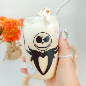 May include: A clear glass with a black and white Jack Skellington design, filled with a light brown beverage topped with whipped cream and cinnamon. A clear glass straw is in the glass.