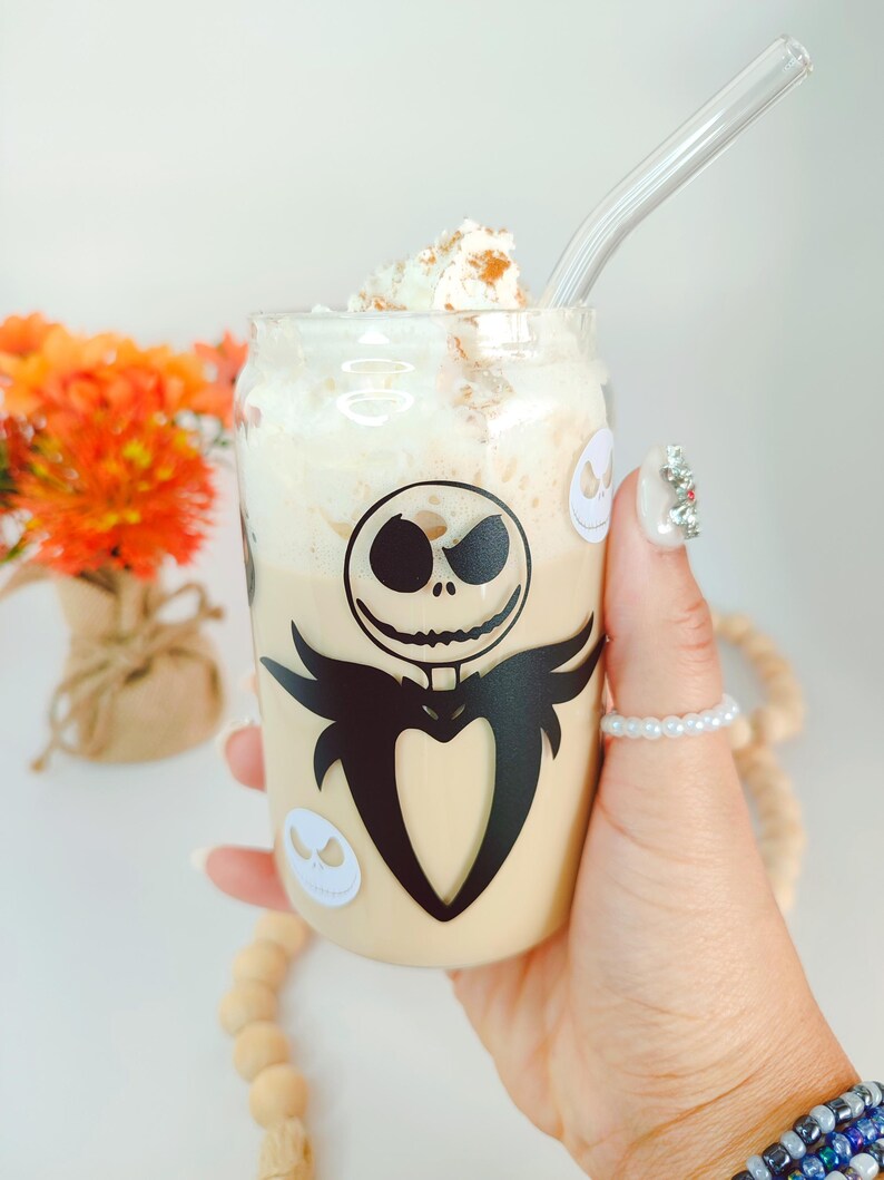 May include: A clear glass with a black and white Jack Skellington design, filled with a light brown beverage topped with whipped cream and cinnamon. A clear glass straw is in the glass.
