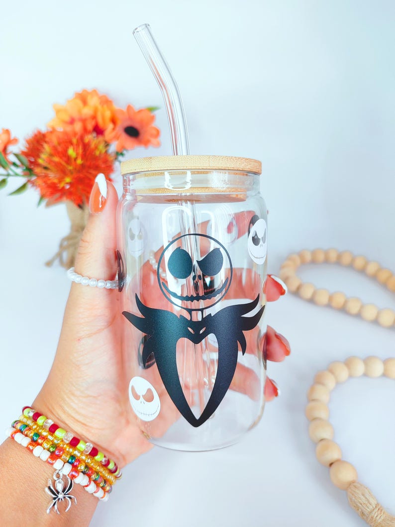 May include: A clear glass jar with a black and white Jack Skellington design and a wooden lid. The jar has a clear glass straw.