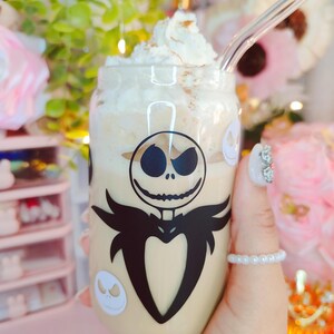 May include: A clear glass mug with a white lid and a black and white Jack Skellington design. The mug is filled with a light brown beverage topped with whipped cream.