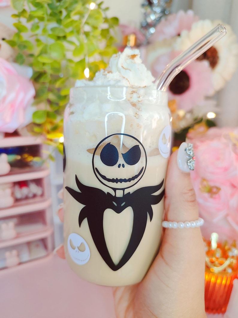 May include: A clear glass mug with a white lid and a black and white Jack Skellington design. The mug is filled with a light brown beverage topped with whipped cream.