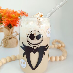 May include: A glass mug with a black and white image of Jack Skellington from The Nightmare Before Christmas. The mug is filled with a light brown beverage topped with whipped cream and a sprinkle of cinnamon. A clear glass straw is sticking out of the mug.