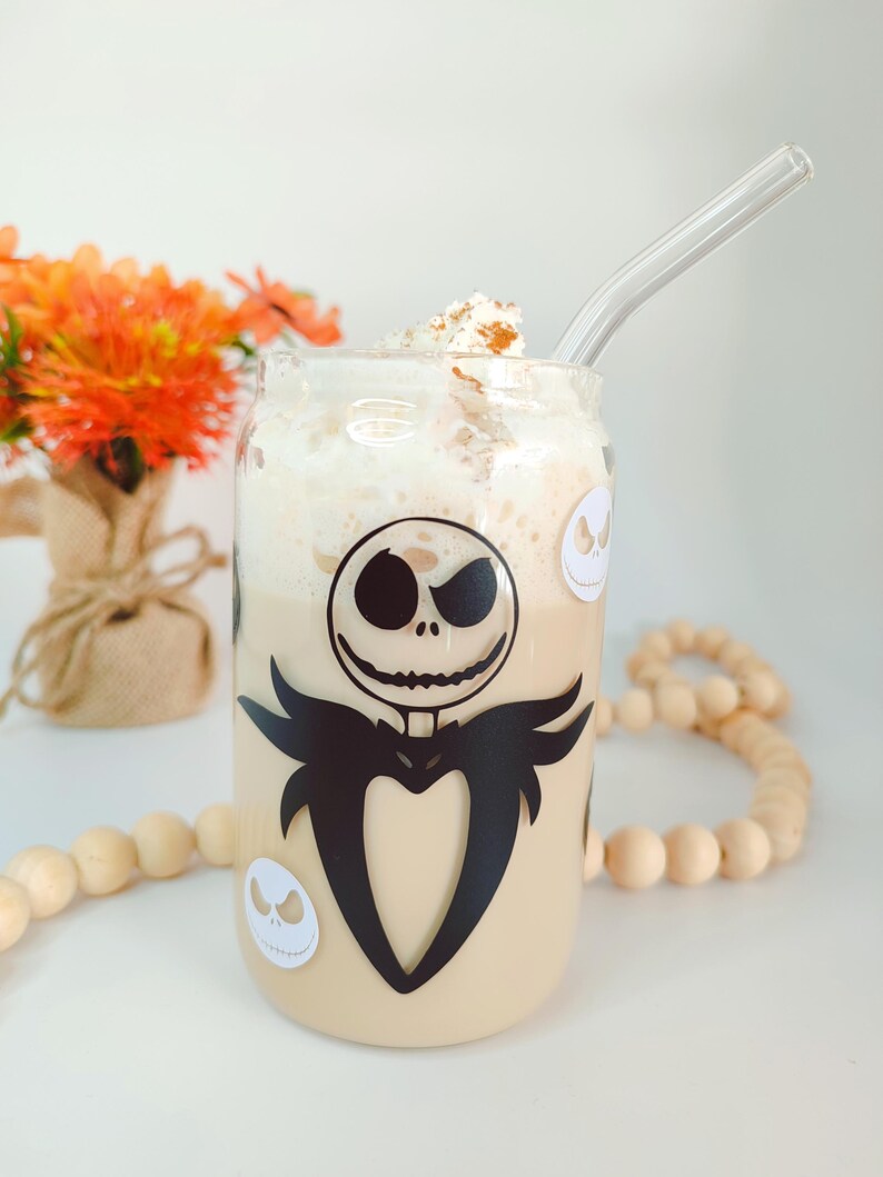 May include: A glass mug with a black and white image of Jack Skellington from The Nightmare Before Christmas. The mug is filled with a light brown beverage topped with whipped cream and a sprinkle of cinnamon. A clear glass straw is sticking out of the mug.