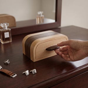 Peut inclure: Une boîte à bijoux en bois au design incurvé, avec un extérieur en bois clair et une bande en bois foncé. La boîte a une poignée ovale foncée et est sur une surface marron foncé. D'autres accessoires sont visibles.