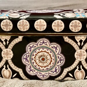 May include: A rectangular black decorative box with a hinged lid. The box is adorned with a repeating floral pattern in white, purple, and teal. The lid features a series of circular designs. A gold-colored band accents the box's midsection.