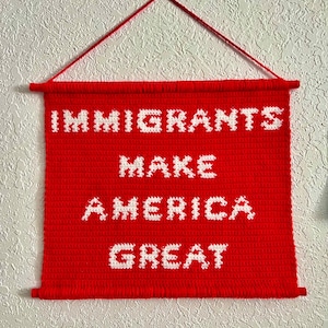 May include: A red crocheted wall hanging with the words "Immigrants Make America Great" in white yarn.