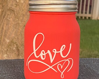 May include: A coral-colored mason jar with a silver lid. The jar has the word "love" written in white cursive with a heart symbol.