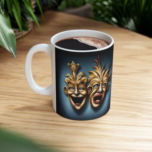 Op de afbeelding: Witte keramische koffiemok met een zwarte handgreep. De mok heeft een kleurrijk beeld van twee gouden theatermaskers op een blauwe achtergrond. De maskers zijn er een lachend en een huilend.