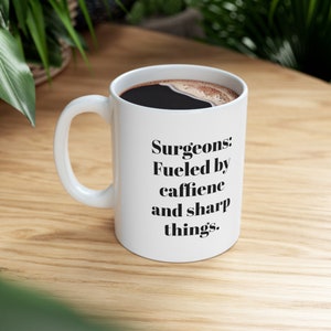May include: White ceramic coffee mug with a black handle. The mug has black text that reads "Surgeons: Fueled by caffeine and sharp things."