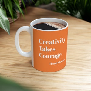 May include: White ceramic mug with an orange band featuring the quote "Creativity Takes Courage" by Henri Matisse in white text. The mug is filled with dark coffee and sits on a wooden surface.