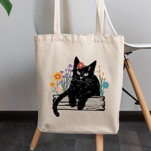 May include: A beige tote bag with a black cat wearing a flower crown and sitting on a wooden plank. The cat is surrounded by colorful flowers.