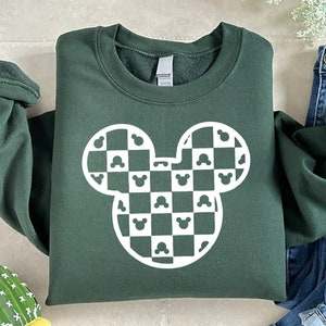 May include: Dark green sweatshirt featuring a white Mickey Mouse head design with a checkered pattern. The design incorporates Mickey Mouse silhouettes within the checks. The sweatshirt is folded, with the sleeves rolled up, and is paired with blue jeans.