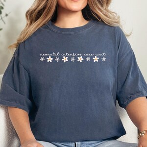 K&ouml;nnte beinhalten: Ein marineblaues T-Shirt mit dem Text "neonatal intensive care unit" und einer Reihe wei&szlig;er Blumen, die auf der Vorderseite aufgedruckt sind. Das Shirt wird von einer Person getragen, die auf einem wei&szlig;en Sofa sitzt.