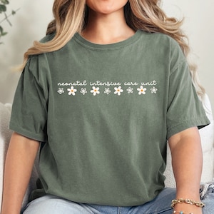 K&ouml;nnte beinhalten: Ein gr&uuml;nes T-Shirt mit dem Text "neonatal intensive care unit" und kleinen wei&szlig;en Blumen, die auf der Vorderseite aufgedruckt sind.