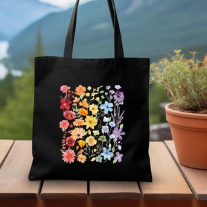 May include: A black tote bag with a rainbow floral design. The flowers are arranged in a rainbow pattern, with red, orange, yellow, green, blue, and purple flowers.
