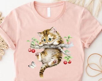 May include: A pink t-shirt featuring a watercolour illustration of a brown and white kitten hanging from a branch with red cherries. A white butterfly is flying near the kitten.