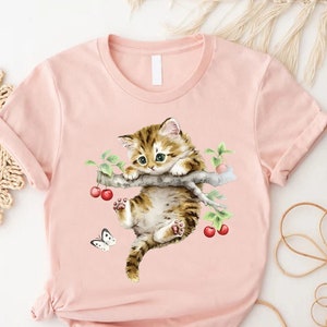 May include: A pink t-shirt with a watercolor illustration of a brown and white kitten hanging from a branch with red cherries. A white butterfly is flying near the kitten.
