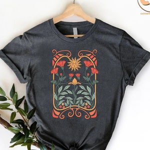 May include: A dark grey t-shirt with a graphic design of a stylized sun with a smiling face surrounded by red flowers and green leaves in an art nouveau style frame.