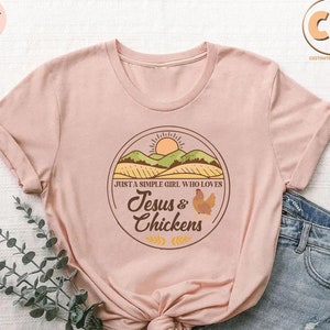 K&ouml;nnte beinhalten: Ein rosa T-Shirt mit einem braunen Kreis-Motiv, das einen Sonnenuntergang &uuml;ber einem gr&uuml;nen Feld, ein Huhn und den Text "Just a simple girl who loves Jesus & Chickens" zeigt.