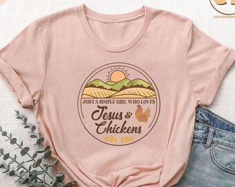 May include: A pink t-shirt with a brown circle graphic featuring a sunset over a field, a chicken, and the text "Just a simple girl who loves Jesus & Chickens".
