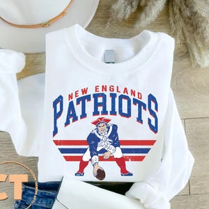 May include: White crew neck sweatshirt featuring the words "NEW ENGLAND PATRIOTS" in blue and red block letters. Below the text is a graphic of a colonial-era man in a blue coat and red pants holding a football. The sweatshirt is folded.