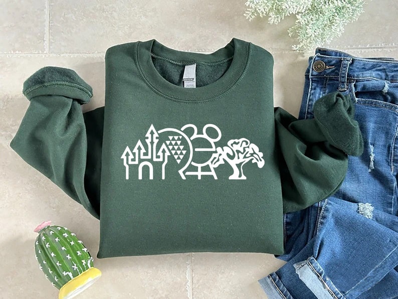 K&ouml;nnte beinhalten: Ein dunkelgr&uuml;ner Pullover mit einer wei&szlig;en Grafik eines Schlosses, einer Kugel mit einem Muster, einem Mickey-Maus-Kopf und einem Baum. Der Pullover ist gefaltet und auf einer gefliesten Oberfl&auml;che abgelegt.