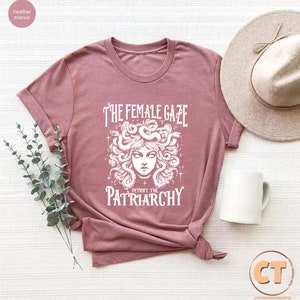 K&ouml;nnte beinhalten: Ein T-Shirt in der Farbe Heide-Mauve mit einem wei&szlig;en Motiv von Medusa mit Schlangen im Haar und dem Text "The Female Gaze Petrified the Patriarchy".