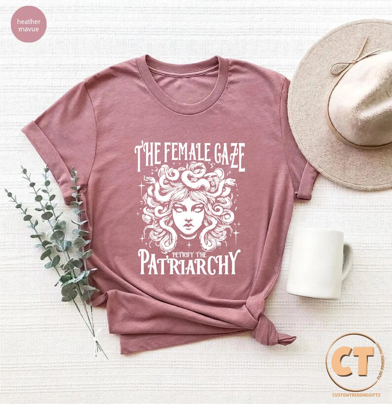 K&ouml;nnte beinhalten: Ein T-Shirt in der Farbe Heide-Mauve mit einem wei&szlig;en Motiv von Medusa mit Schlangen im Haar und dem Text "The Female Gaze Petrified the Patriarchy".