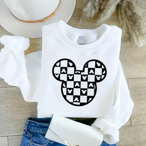 May include: White sweatshirt featuring a black and white checkered Mickey Mouse head design. The sweatshirt is long-sleeved and has a crew neck. The design incorporates Mickey Mouse silhouettes within the checkered pattern. The sweatshirt is folded on a wooden surface.