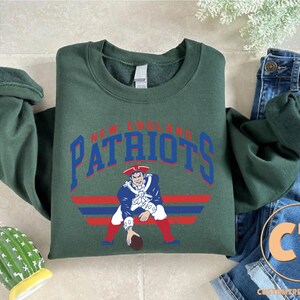 May include: A dark green long-sleeve crewneck sweatshirt featuring the text NEW ENGLAND PATRIOTS in blue arched lettering above a graphic of a football player in a blue, white, and red uniform, positioned over horizontal red and blue stripes.