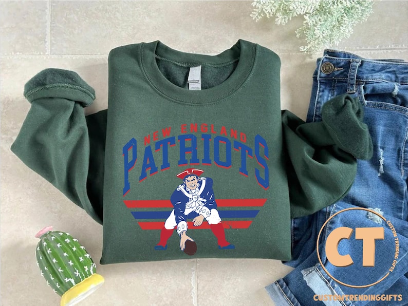 May include: A dark green long-sleeve crewneck sweatshirt featuring the text NEW ENGLAND PATRIOTS in blue arched lettering above a graphic of a football player in a blue, white, and red uniform, positioned over horizontal red and blue stripes.