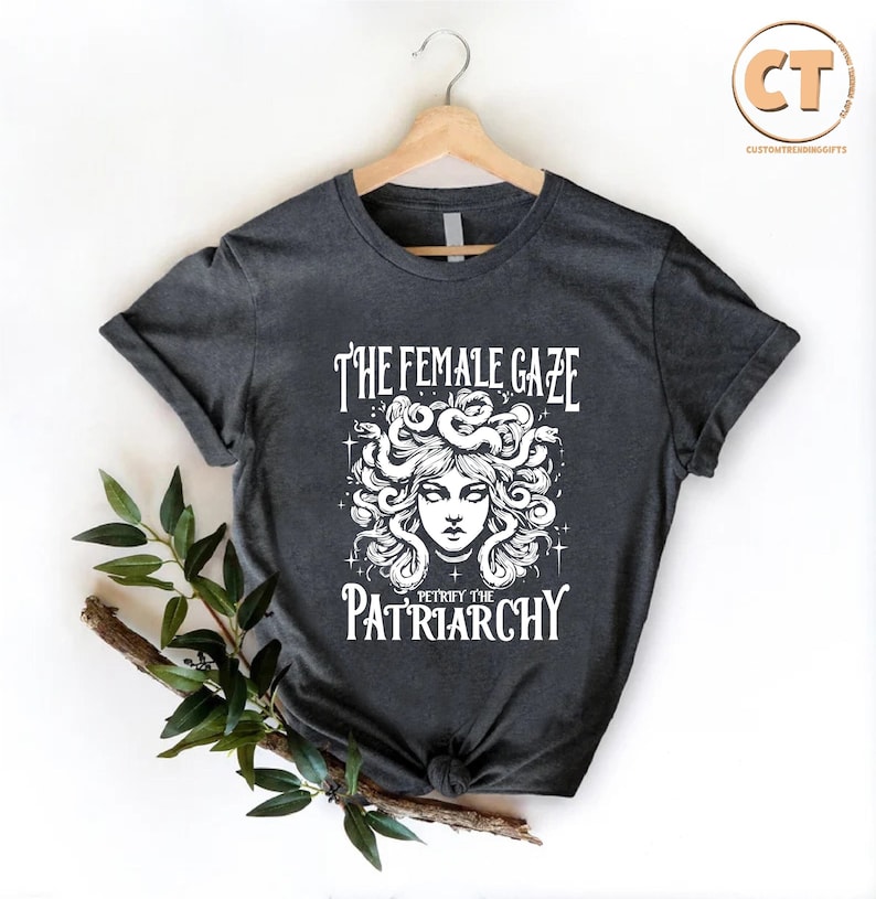 K&ouml;nnte beinhalten: Ein dunkelgraues T-Shirt mit einer wei&szlig;en Grafik einer Frau mit Schlangen als Haaren. Der Text "The Female Gaze" und "Betray the Patriarchy" ist auf dem Shirt aufgedruckt.