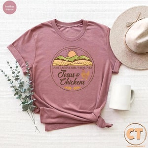 K&ouml;nnte beinhalten: Ein T-Shirt in der Farbe Heide-Mauve mit einem grafischen Design einer Sonne, H&uuml;geln und einem Feld. Der Text auf dem Grafik sagt "Just a simple girl who loves Jesus & Chickens".