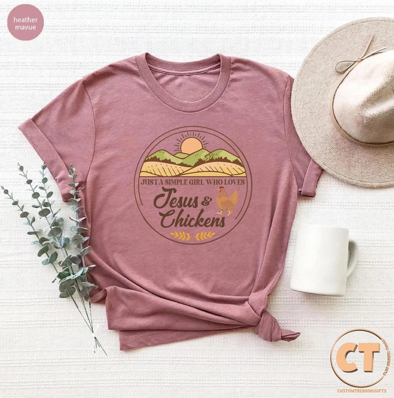 K&ouml;nnte beinhalten: Ein T-Shirt in der Farbe Heide-Mauve mit einem grafischen Design einer Sonne, H&uuml;geln und einem Feld. Der Text auf dem Grafik sagt "Just a simple girl who loves Jesus & Chickens".